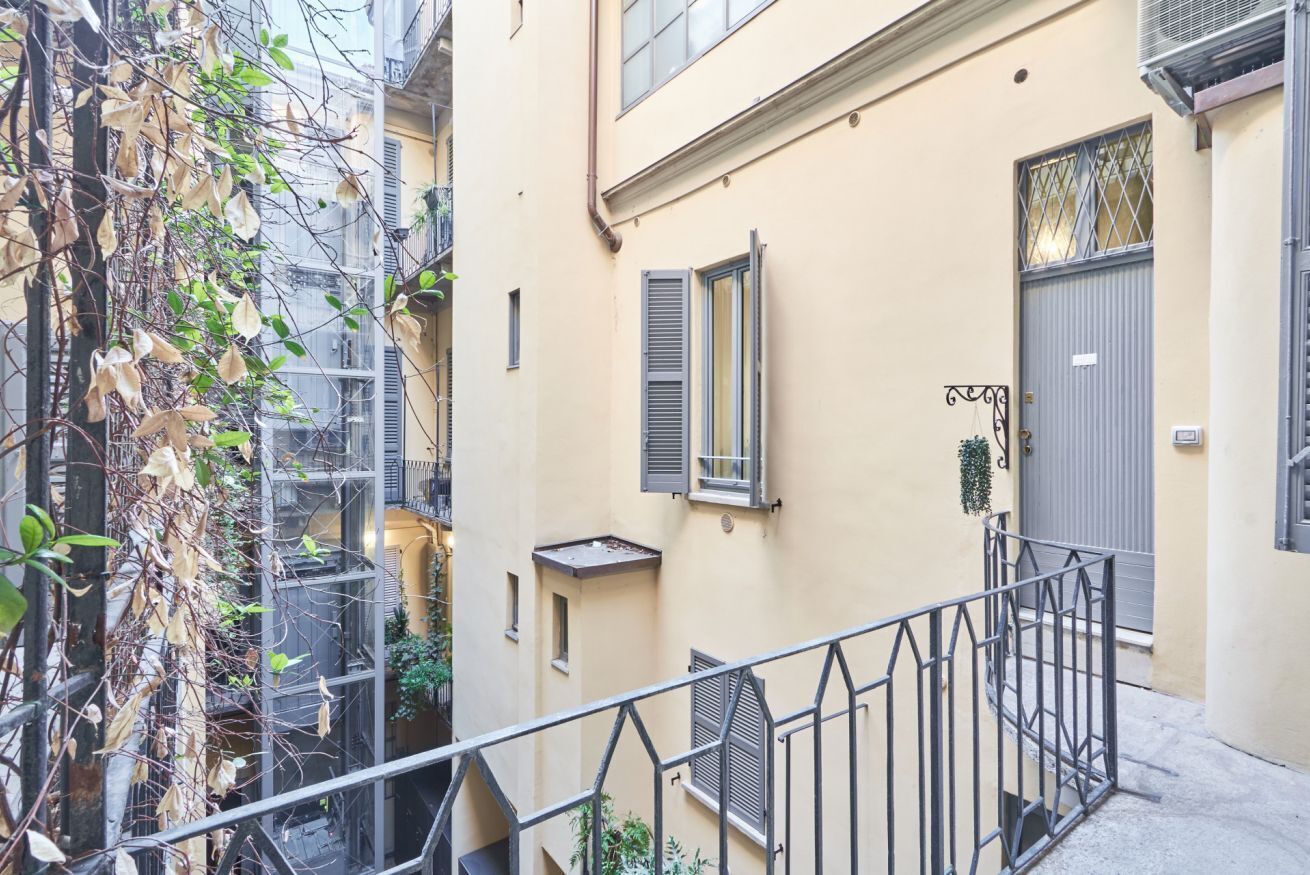 A narrow urban street with a building featuring a balcony, a door, and a window with shutters.