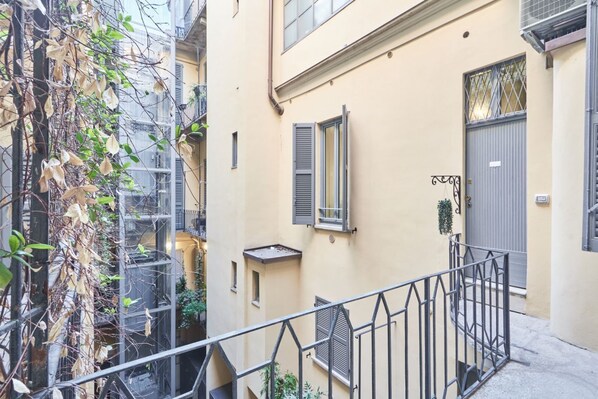 A narrow urban street with a building featuring a balcony, a door, and a window with shutters.