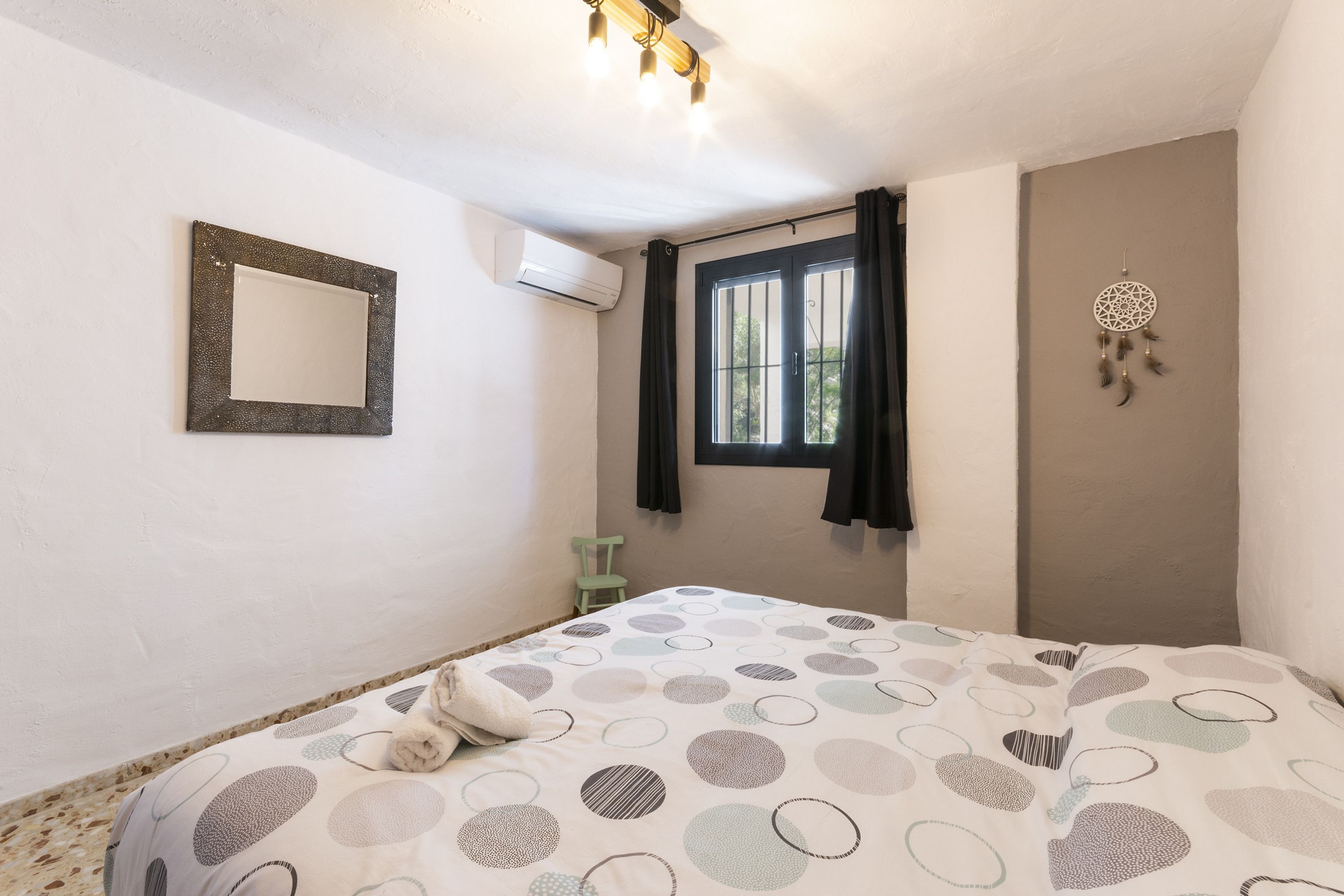 Un dormitorio con cama, ventana con cortinas, aire acondicionado instalado en la pared, un cuadro enmarco y un atrapasueños.