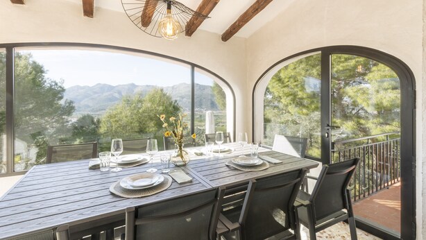 A dining area with a table set for four, large windows offering a mountain view, and a ceiling with exposed wooden beams. - Horizonte in El Capso (Orba)