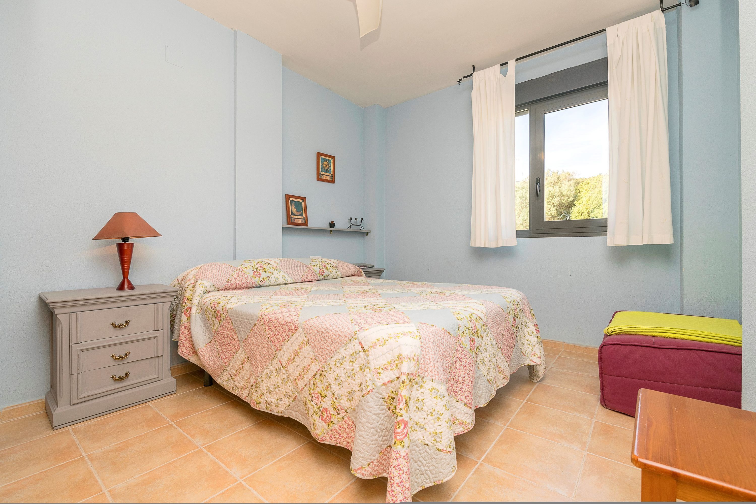 Un dormitorio con cama, mesita de noche, lámpara, ventana con cortinas y una pequeña mesa.