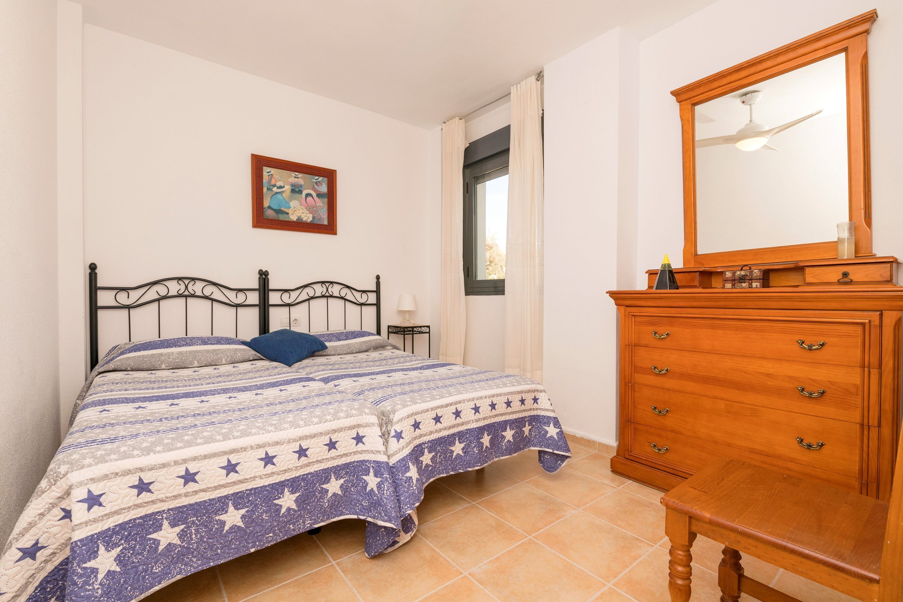 Un dormitorio con cama, cómoda, espejo, ventana con cortinas e imagen colgada en la pared.