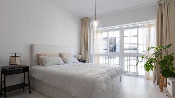 A modern bedroom with a large bed, a bedside table, a hanging light, and a view of the city through a large window. - Jinetes Holidays in M Laga (Málaga)