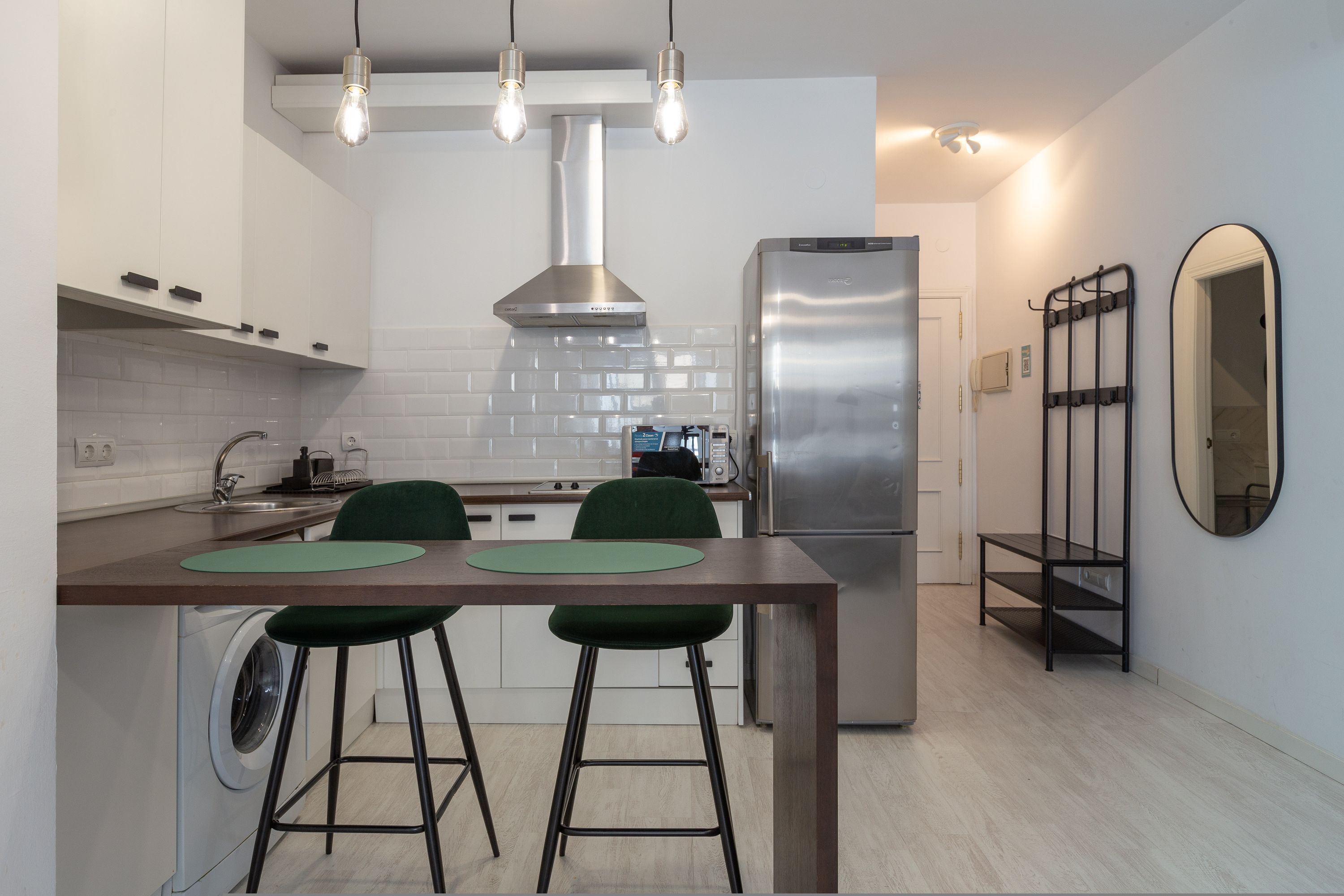 A modern kitchen with a stainless steel refrigerator, a built-in oven, and a sink. There are two green bar stools and a wooden table.