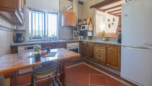 Une cuisine avec des meubles de rangement en bois, un réfrigérateur blanc, une table à manger avec des chaises et une fenêtre avec des stores.
