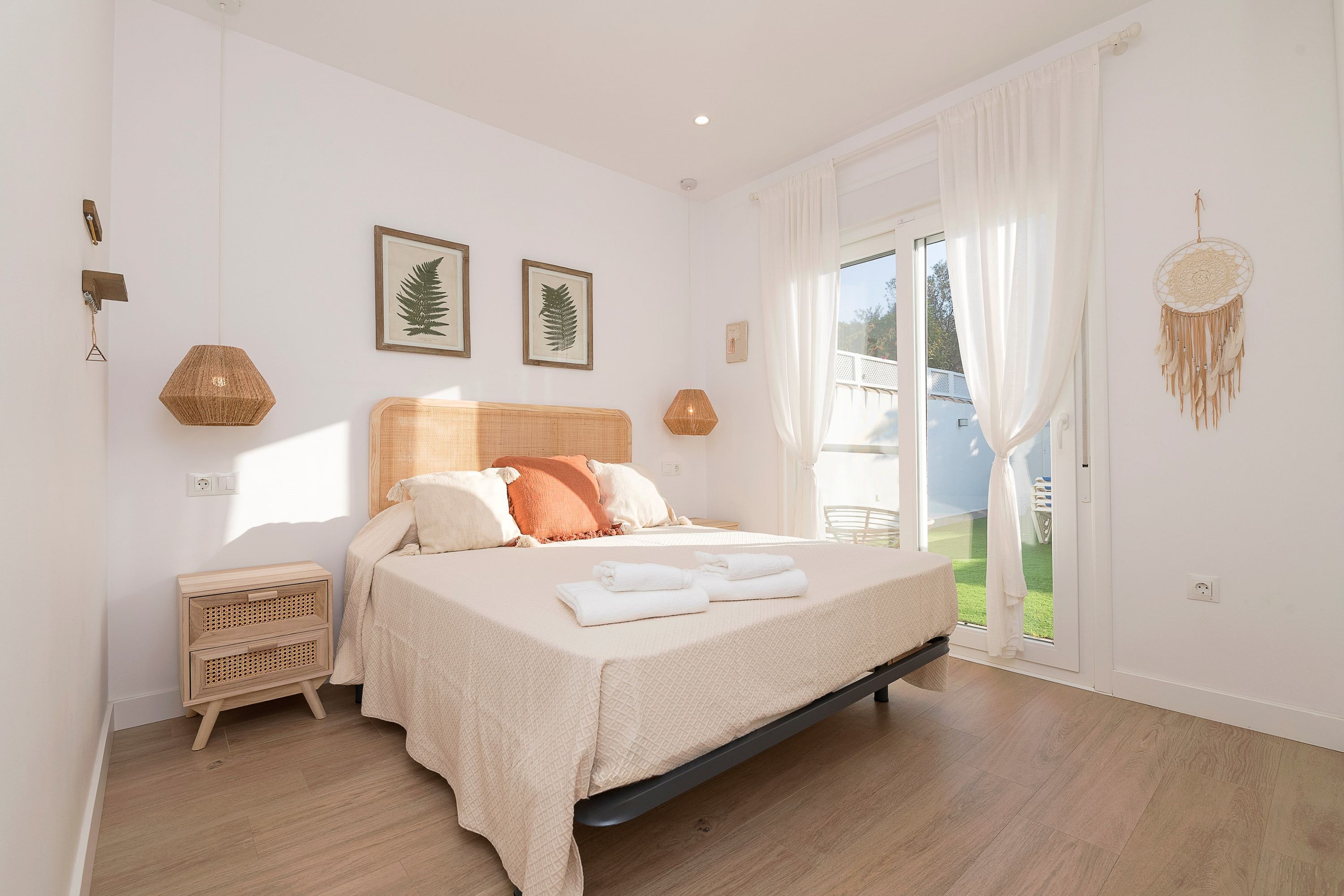 A bedroom with a bed, a bedside table, a window with curtains, and a dreamcatcher on the wall.