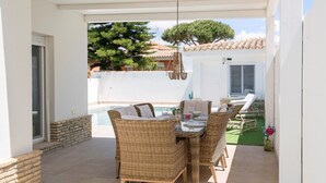 A covered patio with wicker furniture, a hanging light fixture, and a view of a pool and greenery.