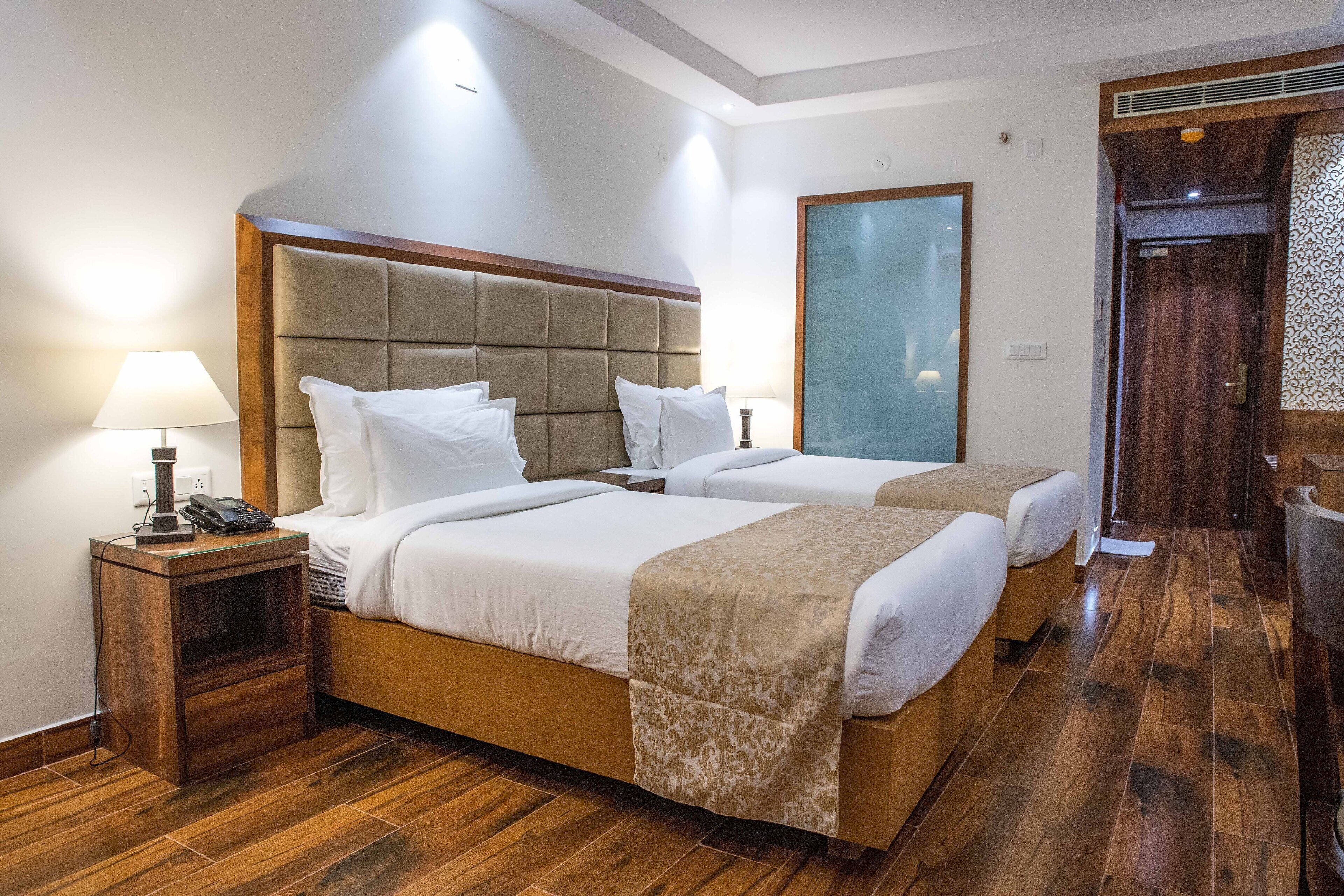 Quarto de hotel com duas camas, piso de madeira, mesa de cabeceira com abajur e um espelho grande.