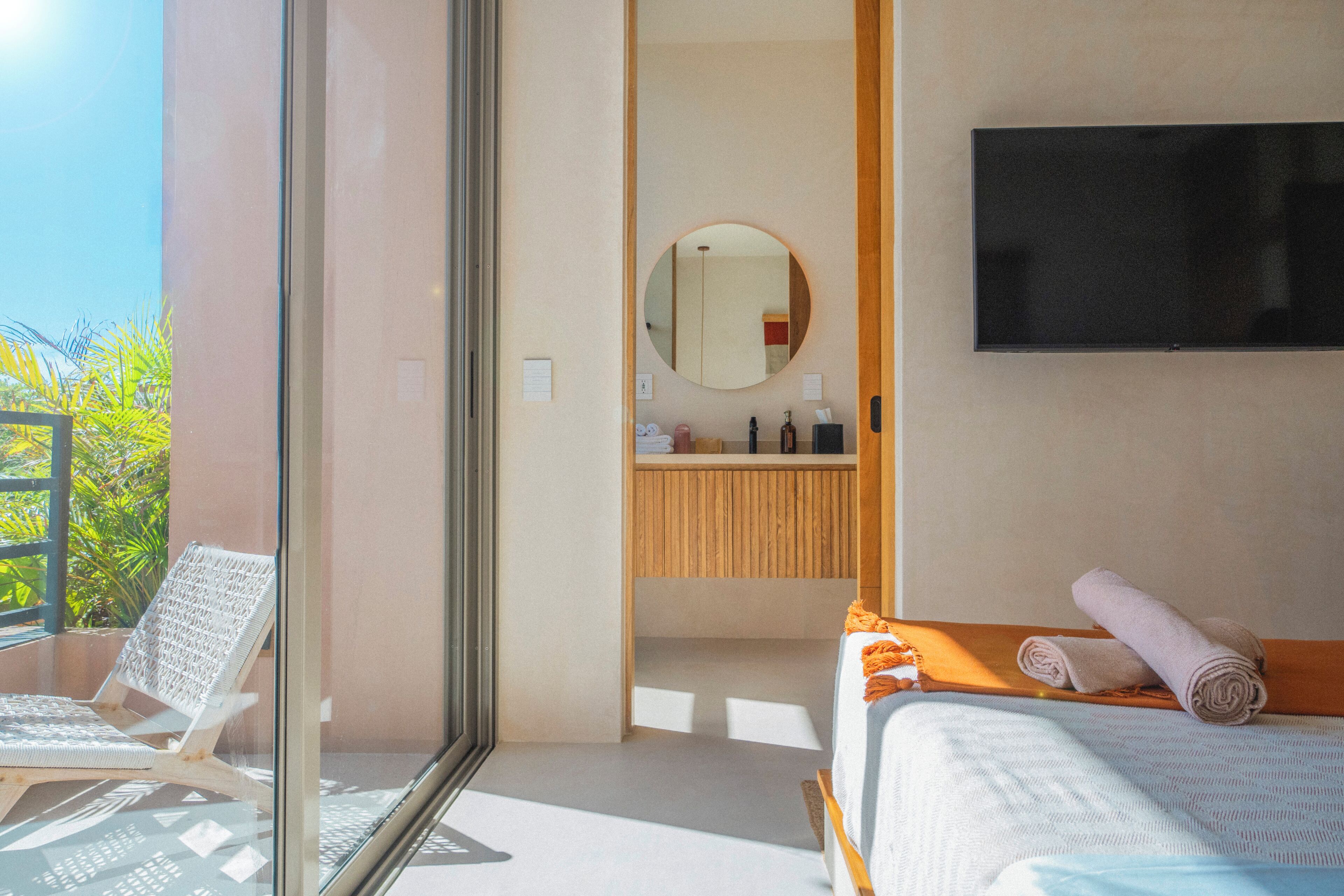 Een slaapkamer met een bed, een televisie, een badkamer met spiegel en een balkon met een stoel.