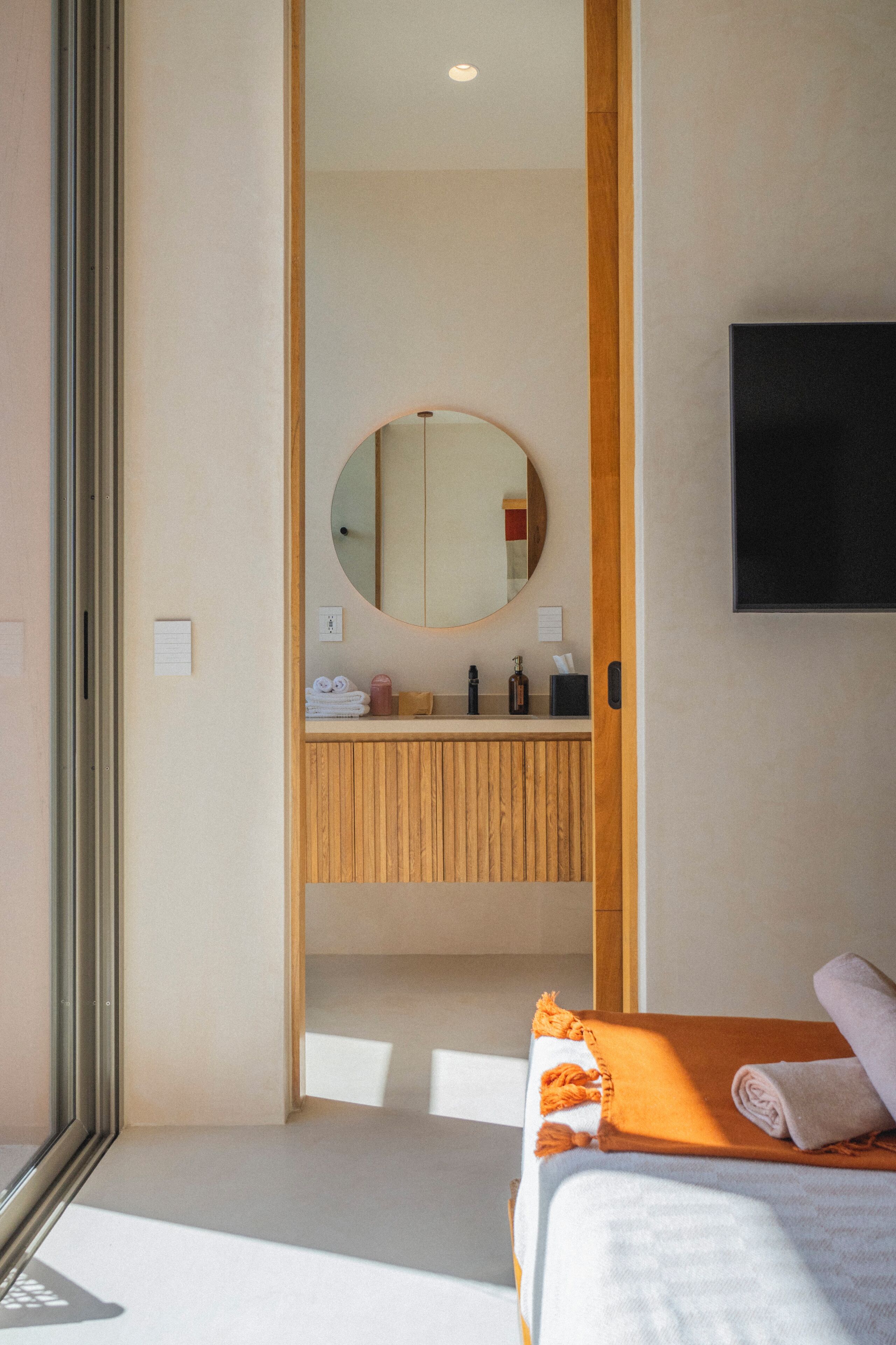 Una camera d'hotel moderna con una toeletta in legno, uno specchio tondo, una TV a schermo piatto e un letto con coperta arancione.