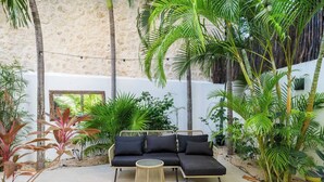 Un área de estar al aire libre acogedora, con un sofá negro y una mesa redonda, rodeada de exuberante vegetación tropical y un muro de piedra.