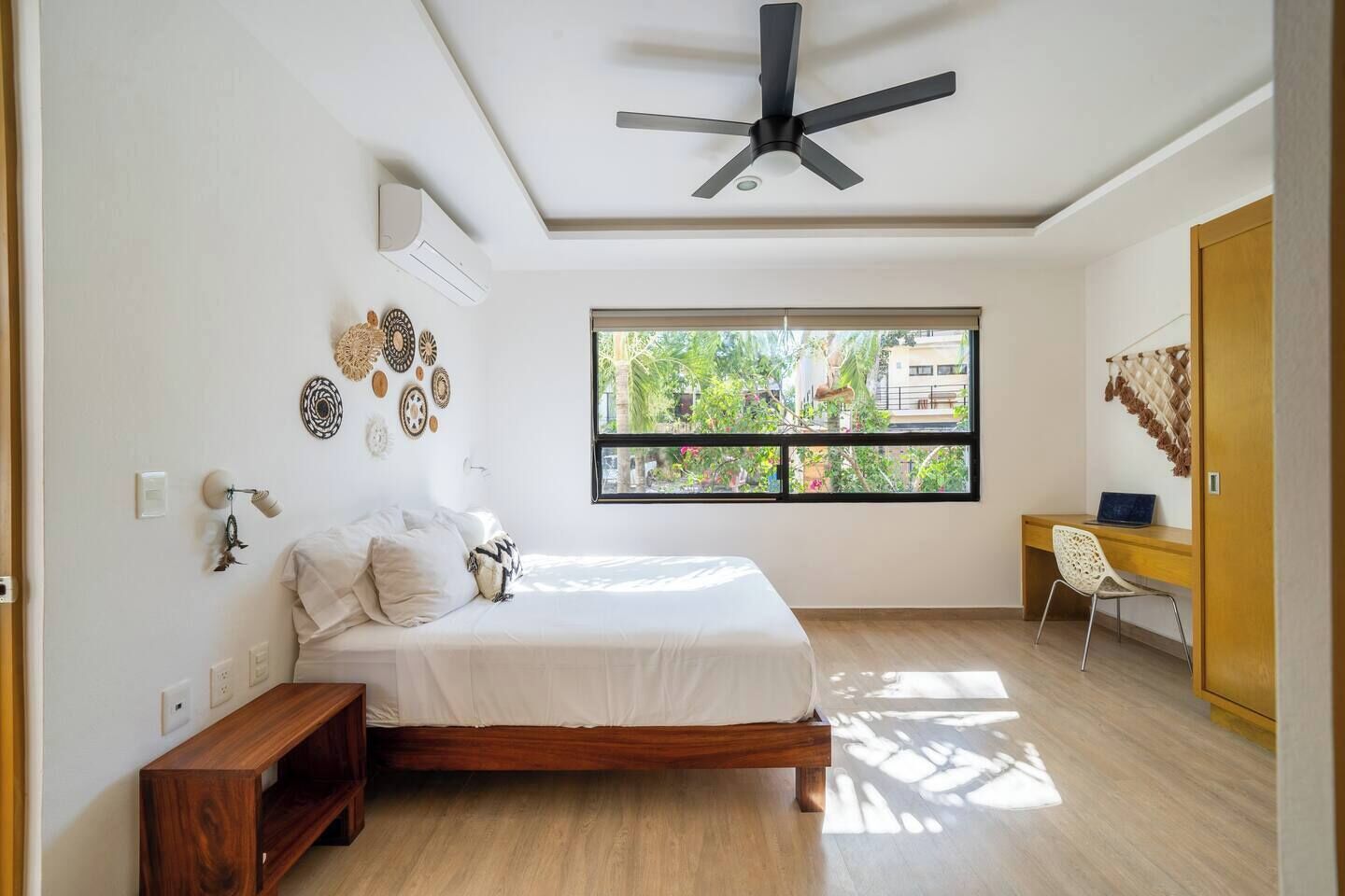 A bedroom with a bed, a desk, a chair, a ceiling fan, and a window with a view of greenery.