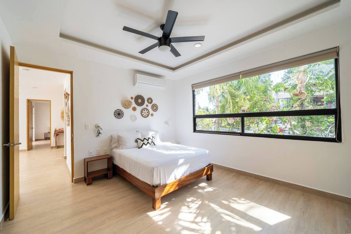 Ein Schlafzimmer mit Bett, Deckenventilator, Fenster mit Blick auf Grünflächen und einer Tür zu einem anderen Raum.