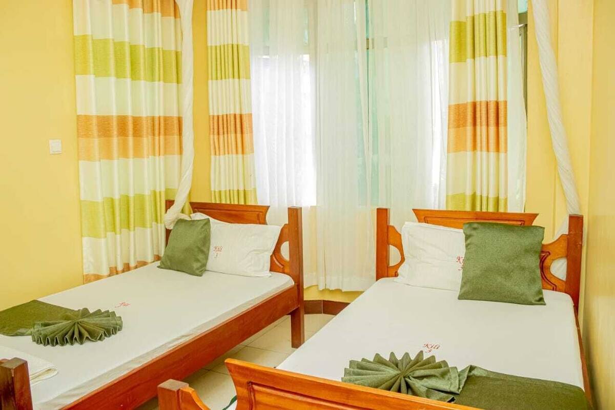 Quarto de hotel com duas camas de solteiro, cada uma com estrutura de madeira e roupa de cama branca. O quarto tem paredes amarelas e cortinas listradas.