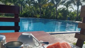 Tavola da pranzo con prodotti per la colazione, tra cui uova strapazzate, anguria e fettine d'arancia, con vista su una piscina e palme.