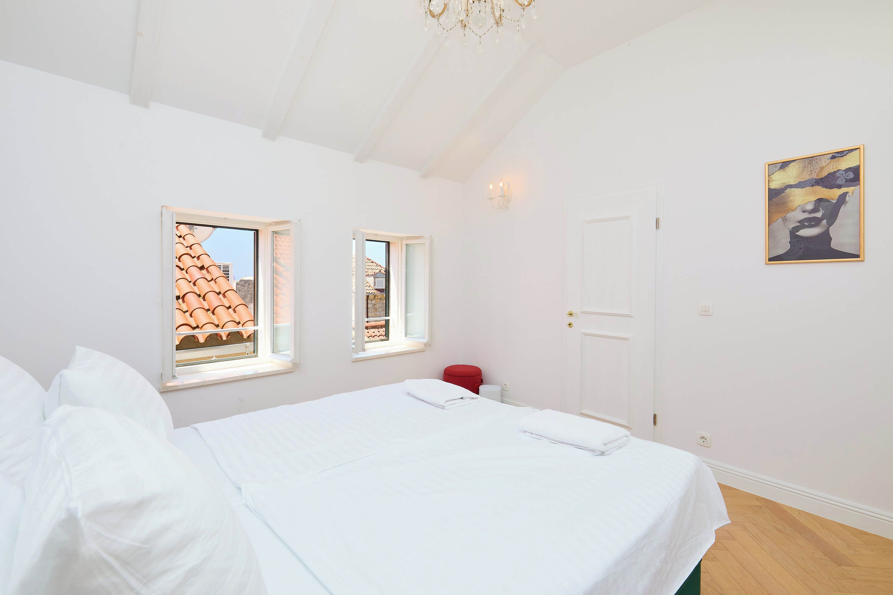 Une chambre à coucher avec un grand lit, deux fenêtres, une porte, un lustre et un tableau au mur.