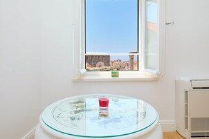 Quarto com mesa de vidro, janela com vista para edifícios e aparelho de ar condicionado.