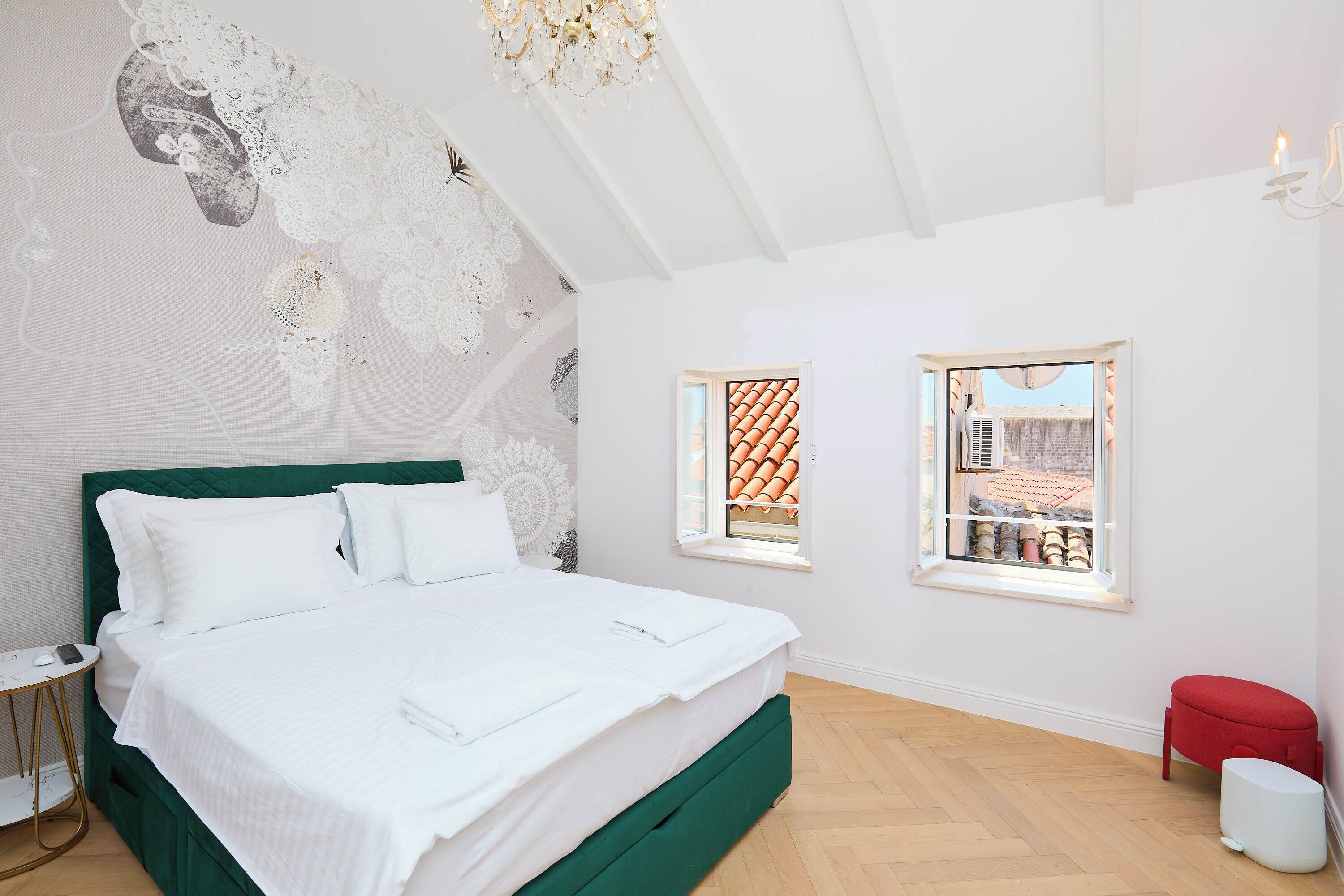 Une chambre avec un grand lit, deux fenêtres, un pouf rouge et une chaise blanche.