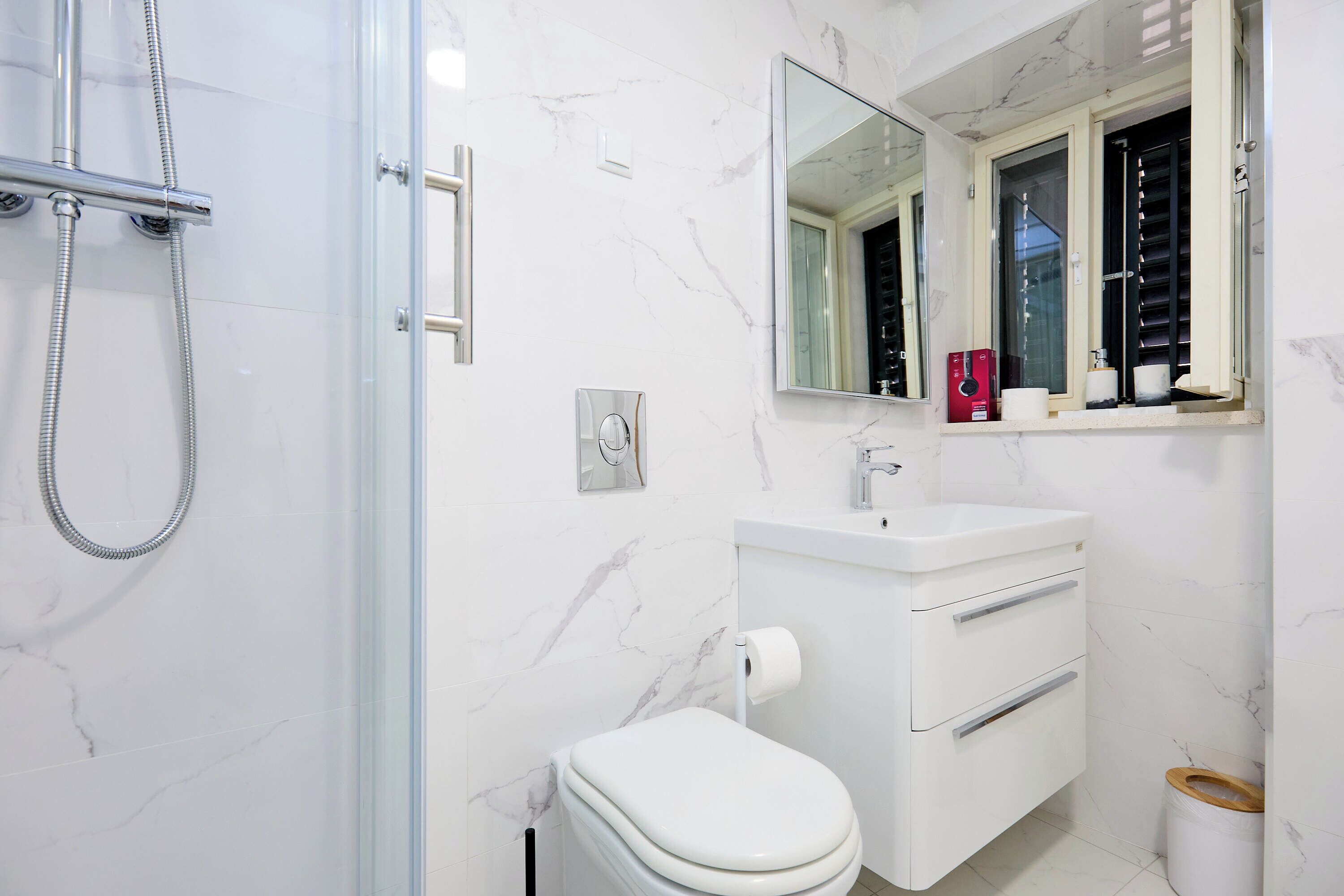 A modern bathroom with a glass-enclosed shower, a white toilet, a vanity with a mirror, and a marble-tiled wall.