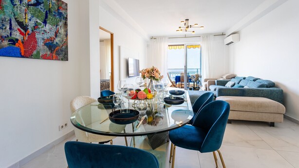 Uma sala de estar moderna com uma mesa de jantar de vidro, cadeiras azuis, um sofá e um quadro abstrato colorido na parede.