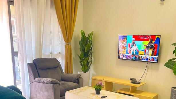 A living room with a teal sofa, a grey armchair, a white coffee table, and a TV mounted on the wall. - Munyonyo 2bedroom Joy Apartments (Kampala)