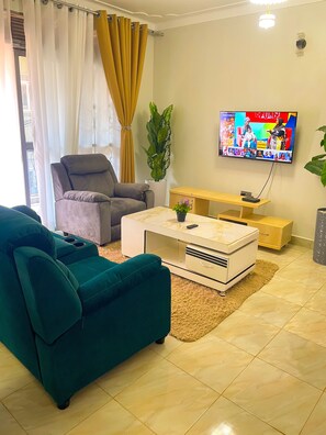 A living room with a teal sofa, a grey armchair, a white coffee table, and a TV mounted on the wall. - Munyonyo 2bedroom Joy Apartments (Kampala)