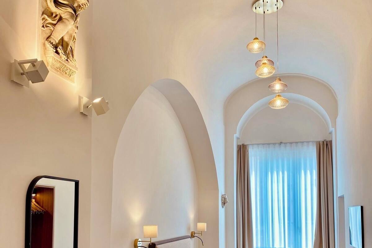 Hotelkamer met een bed, een klein tafeltje, een spiegel en een garderobekast. Er hangen drie hanglampen en een raam met gordijnen.