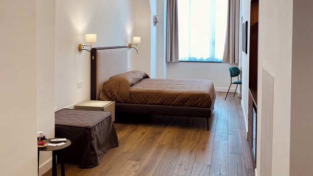 Un dormitorio con cama, mesita de noche, silla, ventana con cortinas y lámpara en el techo.