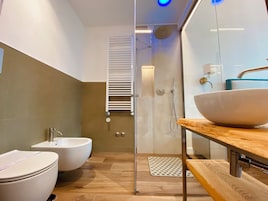 Un baño moderno con bidé, un lavamanos grande de color blanco, cabina de ducha de vidrio y tocador de madera.
