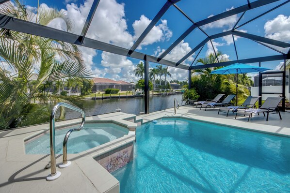 A pool area with a view of a body of water, lounge chairs, and a covered section with a blue umbrella.