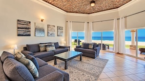 A living room with a large window overlooking a beach, furnished with sofas and a coffee table.