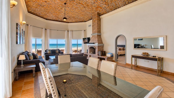 A spacious living area with a glass dining table, comfortable seating, a fireplace, and a view of the beach.