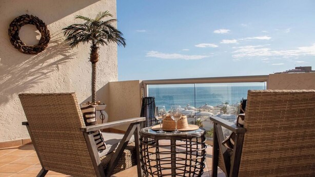 A balcony with wicker chairs, a small round table with a glass, and a view of the ocean.