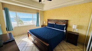 Una habitación de hotel con cama, escritorio con computadora portátil, mesita de noche, ventana con cortinas y vista a la playa.