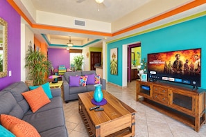 A living room with a sofa, coffee table, and TV. There are plants and a painting on the walls.