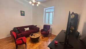 Ein Wohnzimmer mit einem roten Sofa, zwei roten Sesseln, einem Holztisch, einer schwarzen TV-Konsole und einem Holzboden mit Fischgrätmuster.