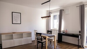 A modern, well-lit room with a wooden dining table, chairs, a white shelving unit, and a desk with a computer. - Apartment New York (Forheim)