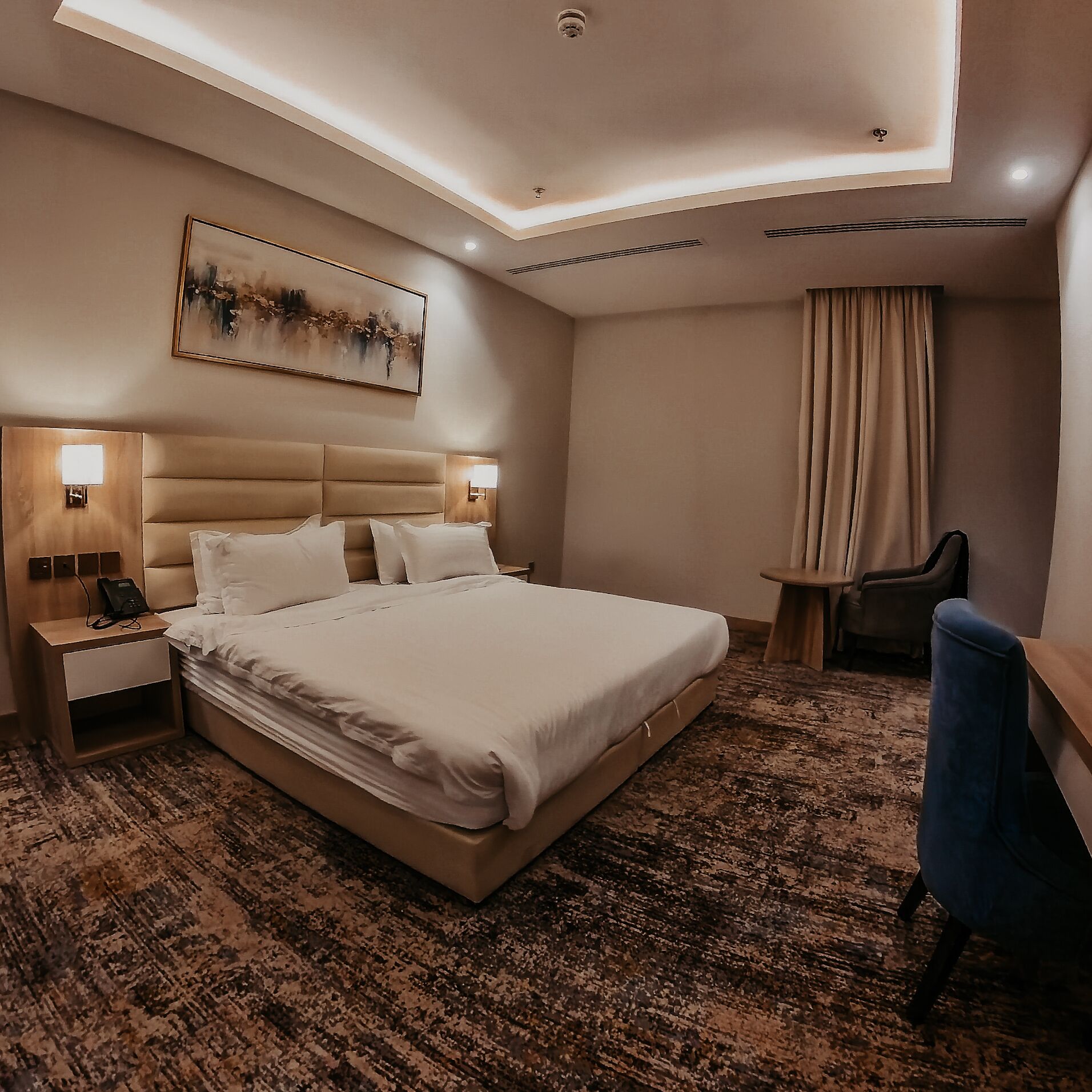 A modern hotel room with a large bed, bedside lamps, a desk, a chair, and a painting on the wall.
