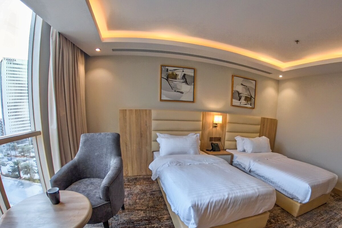 A hotel room with two beds, a small table, a chair, and a large window.