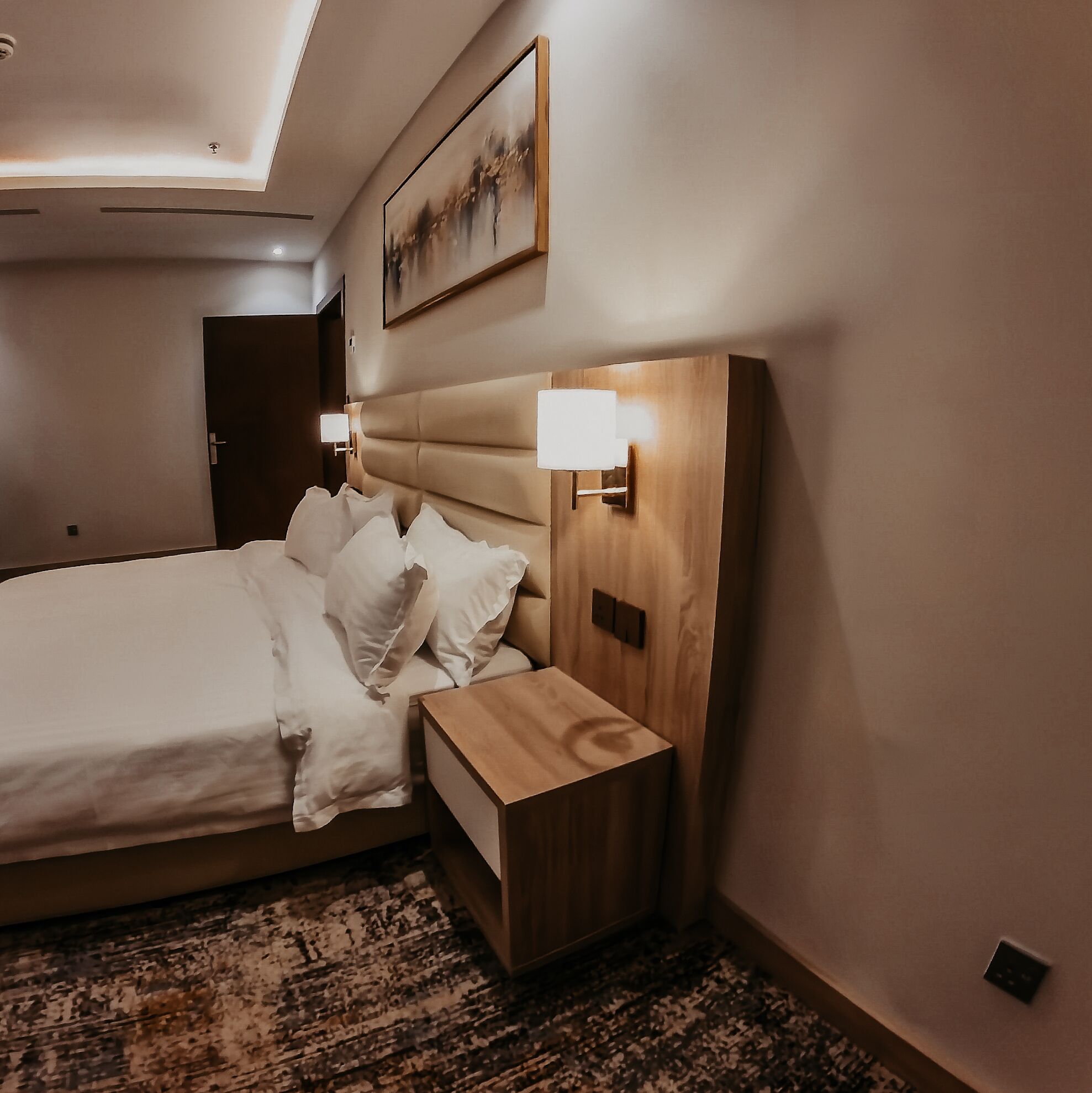 A hotel room with a bed, bedside tables, a headboard, a wall-mounted light, and a painting.