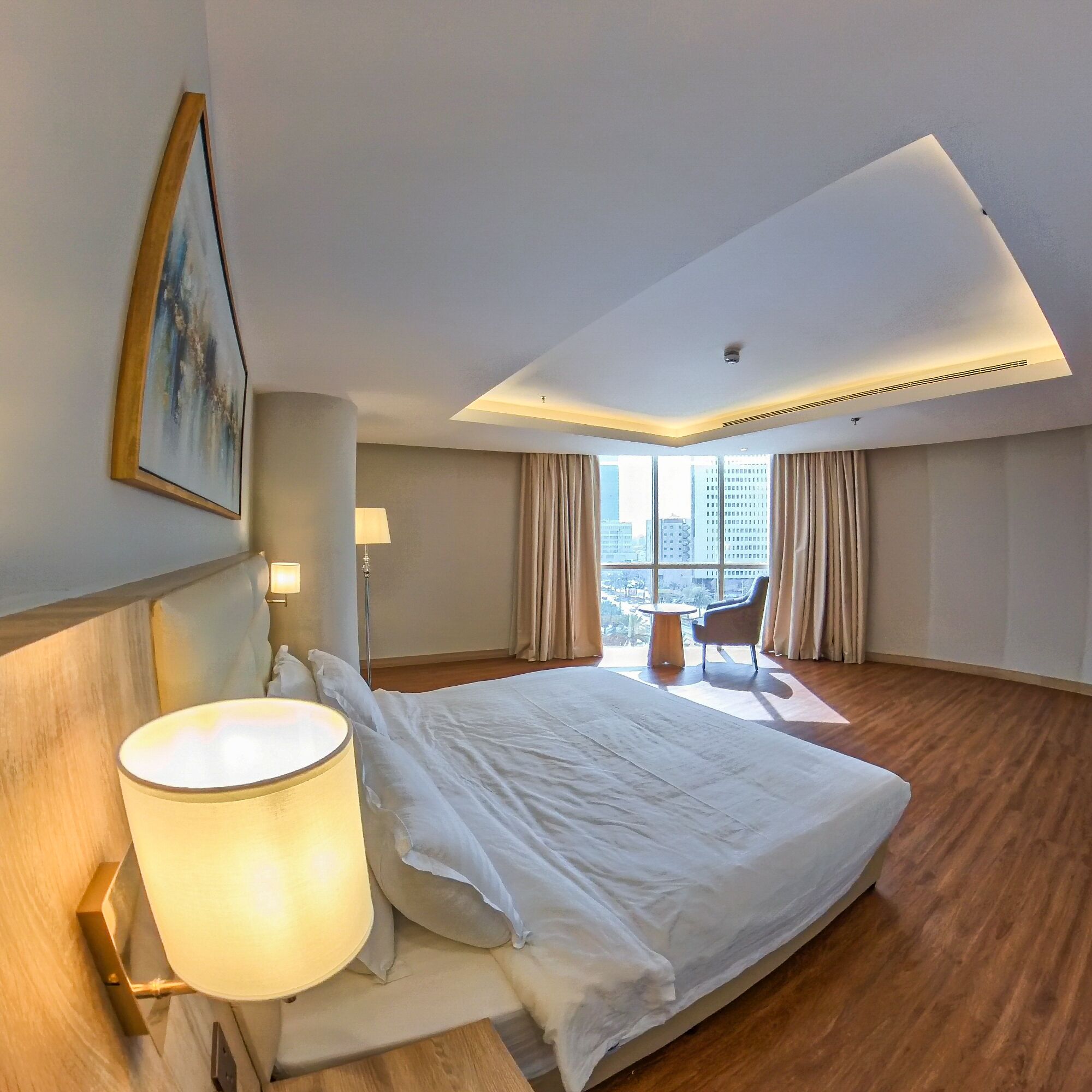 A hotel room with a large bed, a bedside lamp, a chair, a view of the city through large windows, and a ceiling light.