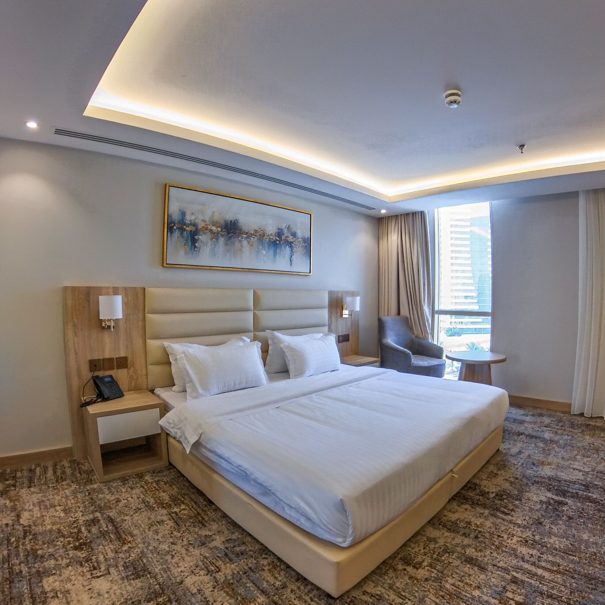 A hotel room with a large bed, bedside tables, a chair, a small table, and a painting on the wall.
