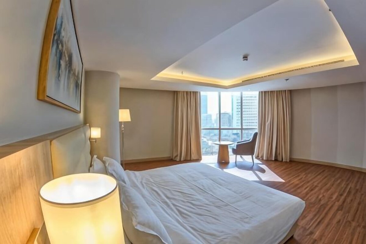 Une chambre d’hôtel avec un grand lit, un bureau, une chaise, une lampe et une vue sur la ville par la fenêtre.