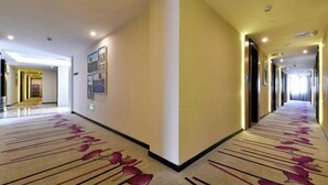 A hotel corridor with a carpet featuring a floral pattern, a light-colored wall with framed pictures, and a ceiling with recessed lighting. - Lavande Hotel (Qingyuan City Square Feilai Lake Branch) (Qingyuan)