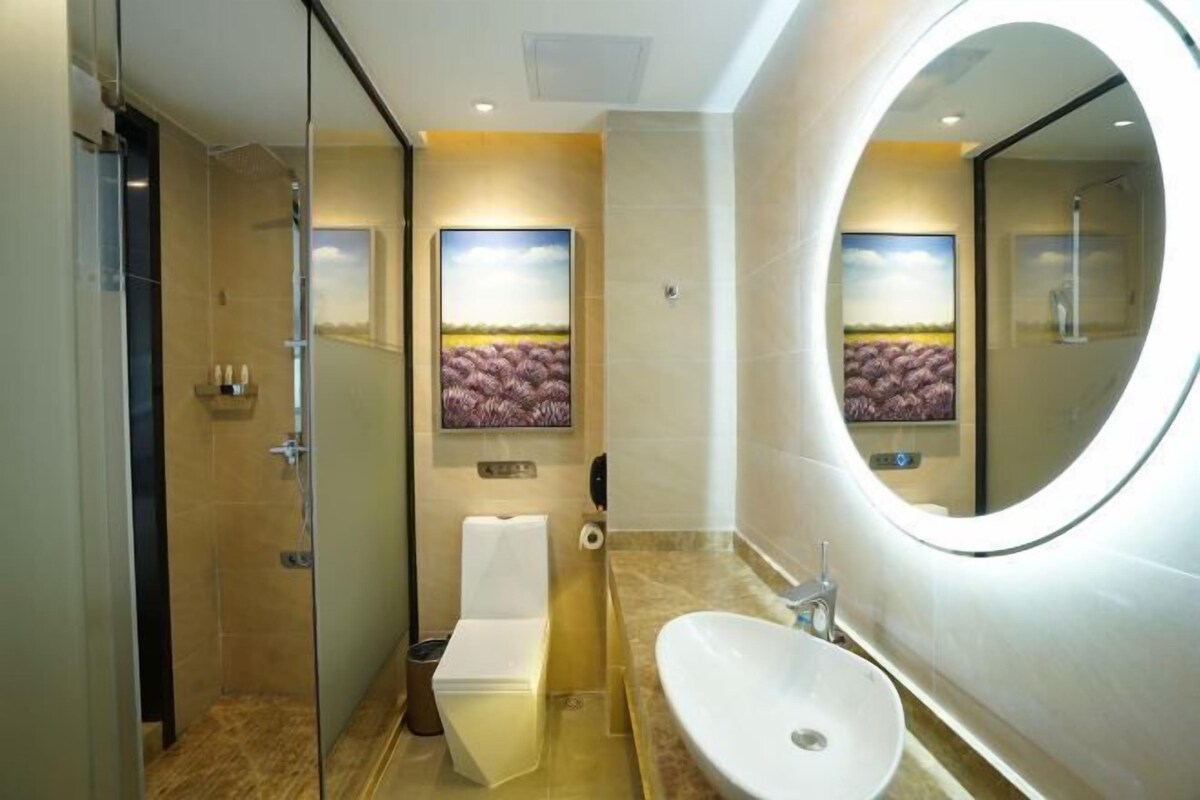 A modern bathroom with a glass shower enclosure, a toilet, a sink with a large mirror, and a framed artwork on the wall.