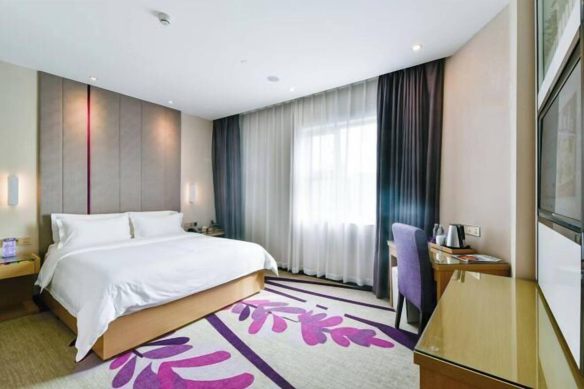 A hotel room with a bed, a desk, a chair, a window with curtains, and a wall-mounted light fixture.