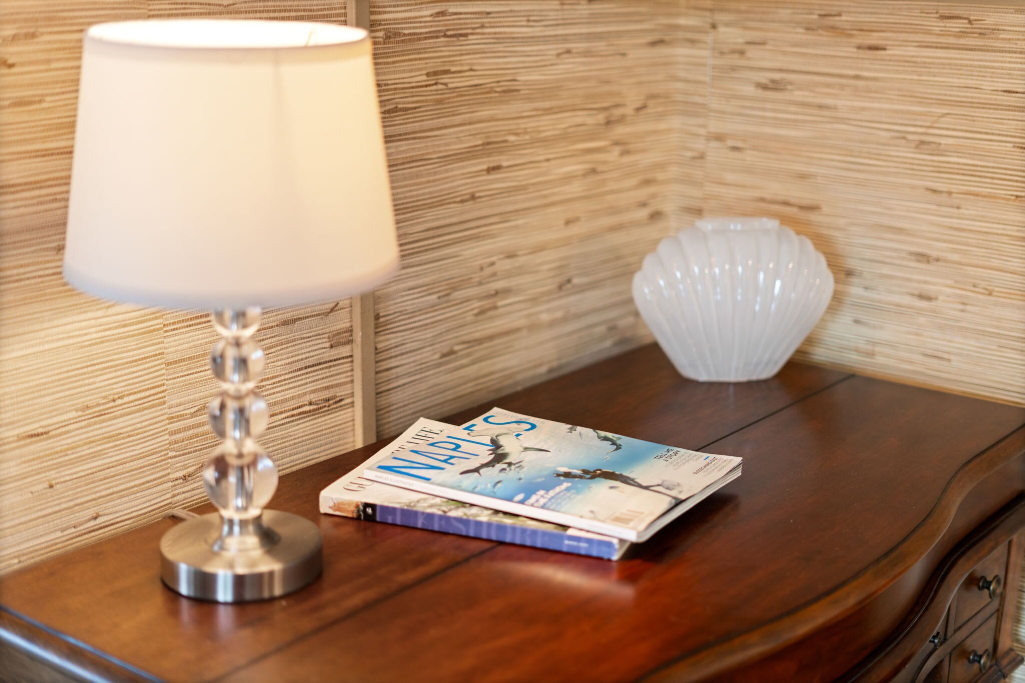 Un comodino in legno con una lampada, un libro intitolato "The Nap of the Times" e un vaso a forma di conchiglia bianca.