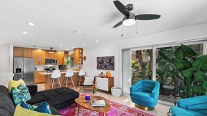 A modern living room with a sofa, coffee table, and blue armchairs. A kitchen with wooden cabinets and a microwave is visible in the background.