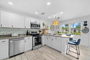 Una cocina moderna con armarios blancos, electrodomésticos de acero inoxidable, una isla grande con taburetes de bar y vistas a un jardín tropical.