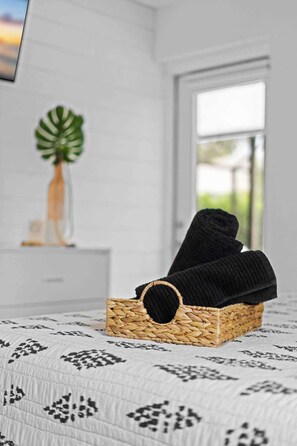 Un lit recouvert d’une couvre-lit à motifs, un panier en osier contenant un objet noir, et une fenêtre donnant sur un paysage verdoyant.